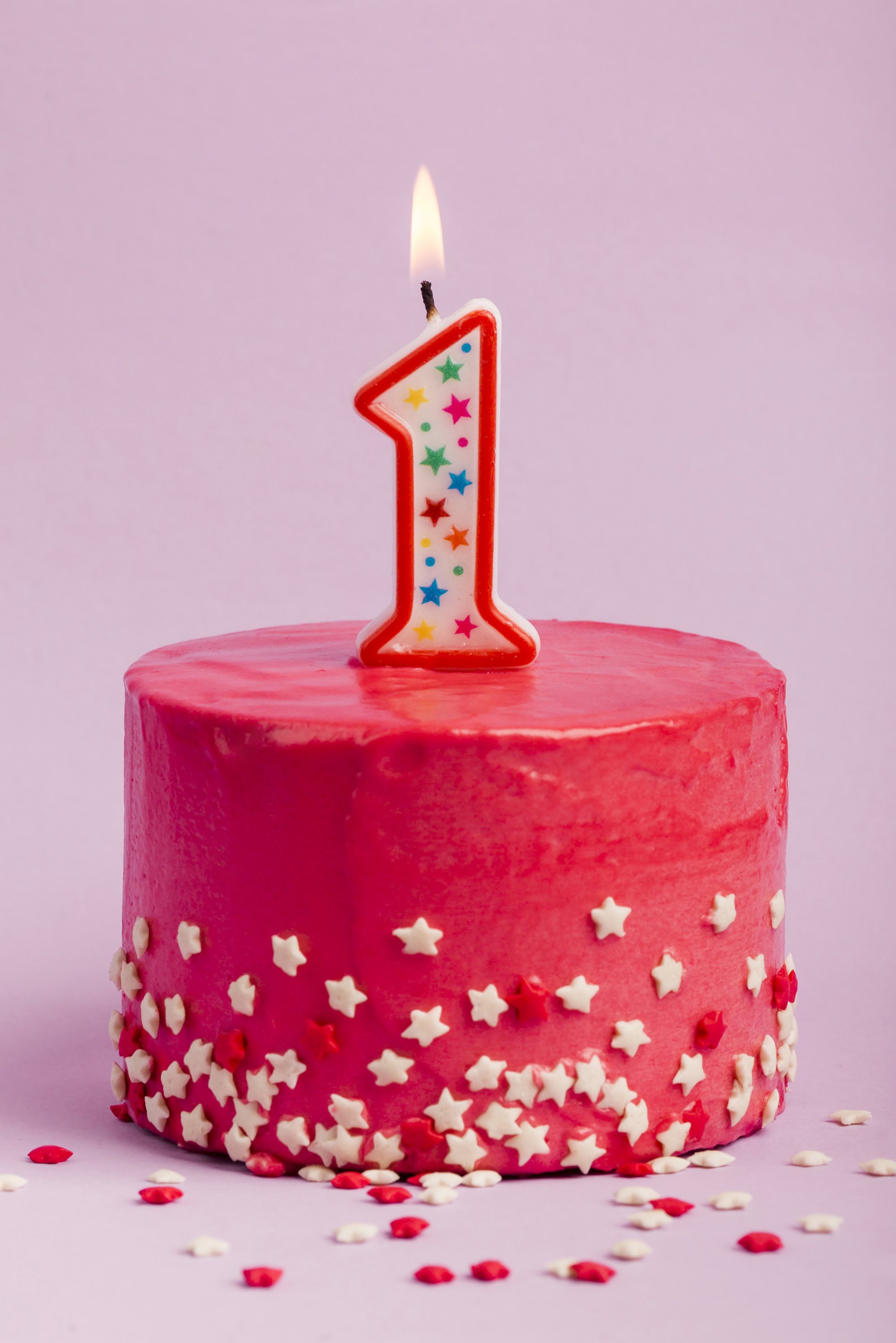 lighted-number-one-candle-on-red-cake-with-star-sprinkles-against-purple-backdrop.jpg