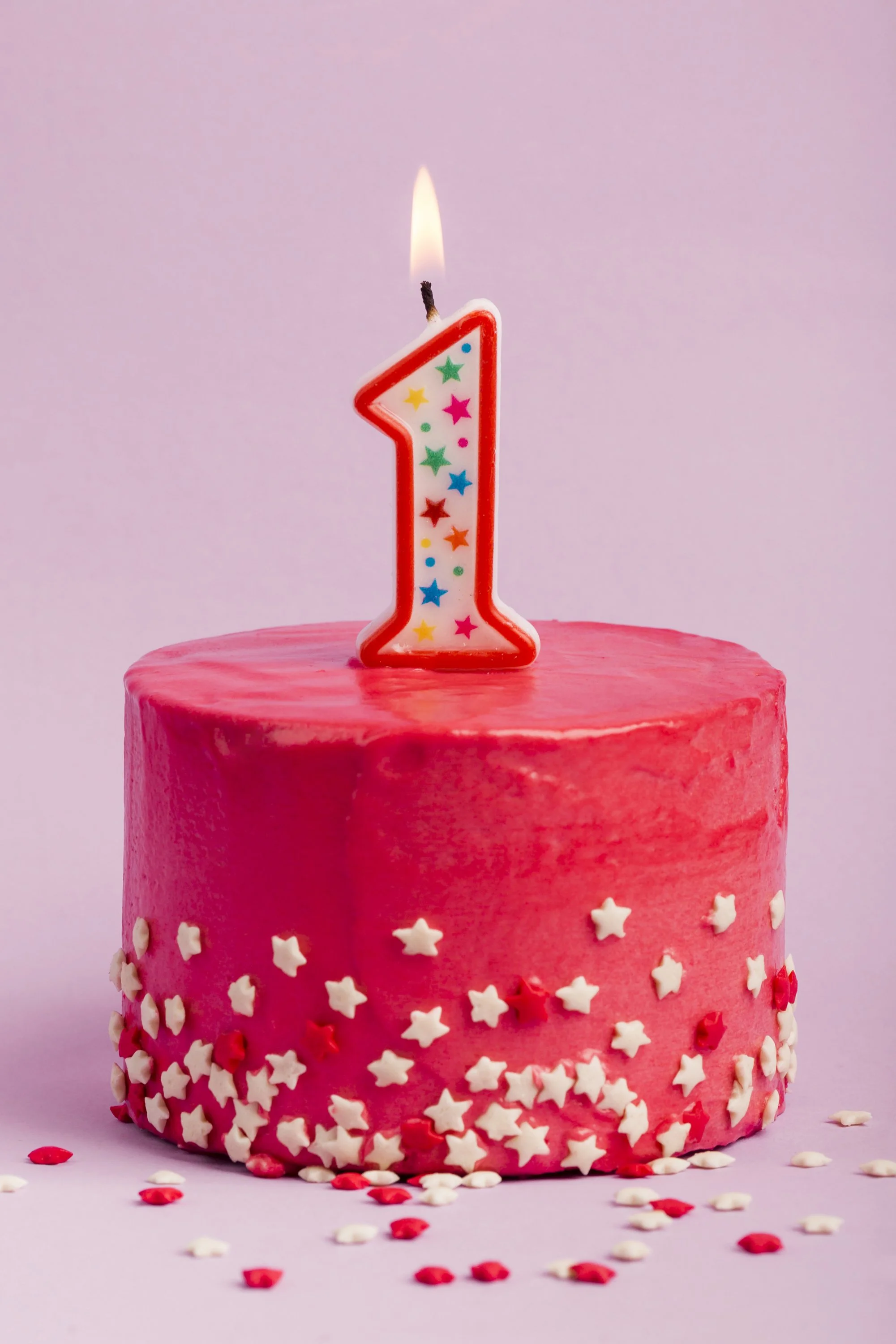 lighted-number-one-candle-on-red-cake-with-star-sprinkles-against-purple-backdrop.webp